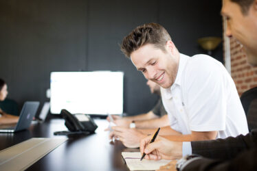 attachment-5613eea9e4b052adbca11866 A man writing in a notebook while another man looks on