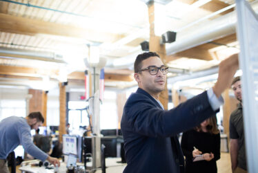 attachment-5613ee00e4b0aab360e5d34a A man writing on a white board while two others look on