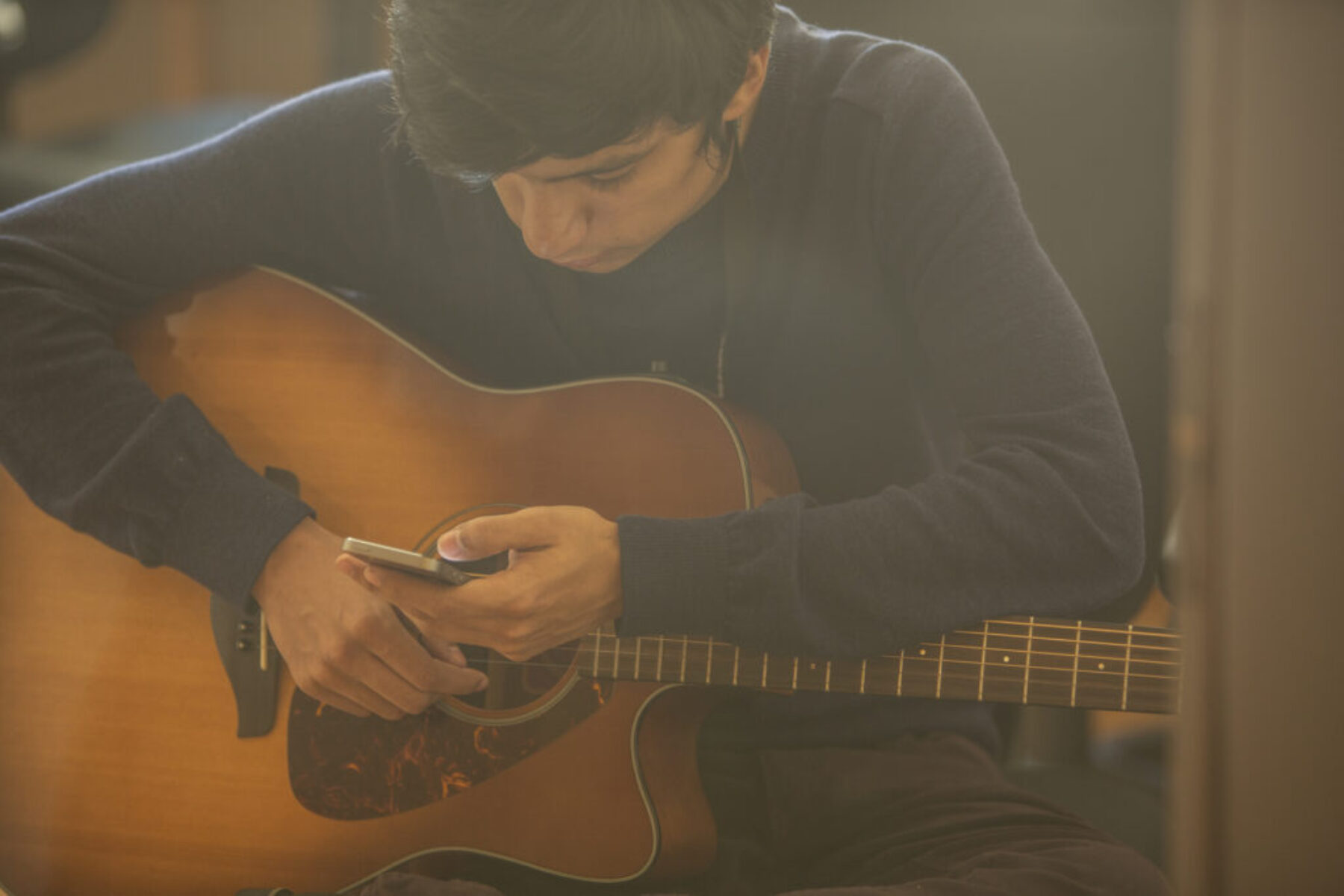 A man holding a guitar while looking down at his phone