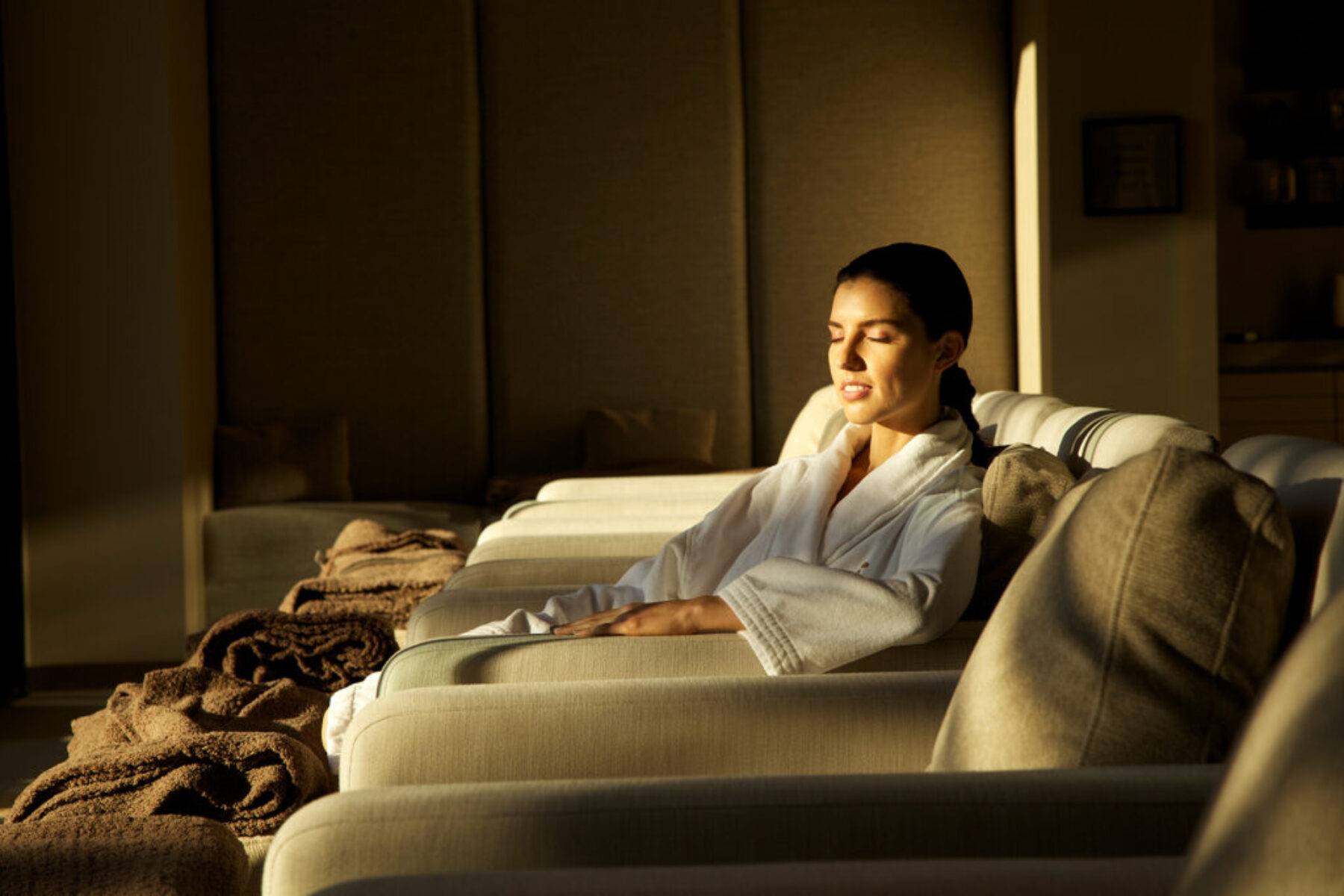 A woman relaxing on a seat indoors with her eyes closed and sun shining on her