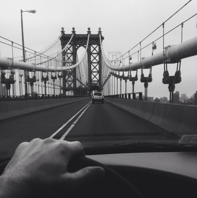 Black and white image of a hand on a steering wheel driving over a bridge in Ney York City