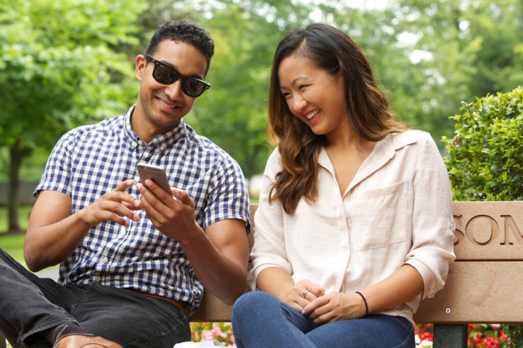 Young man and woman sitting on a park bench smiling and looking at a smartphone together.