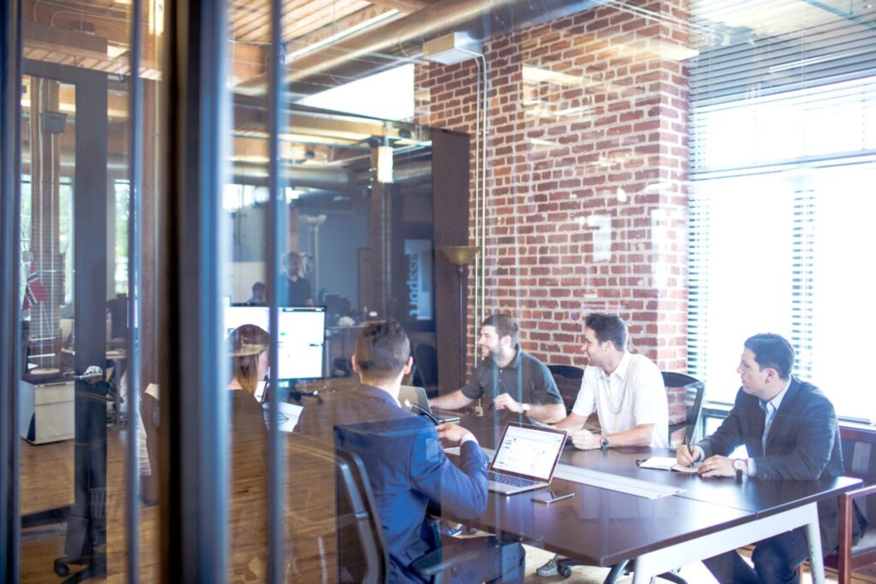 Corporate team meeting in a modern office environment with brick walls, glass conference room, and collaborative workspace.
