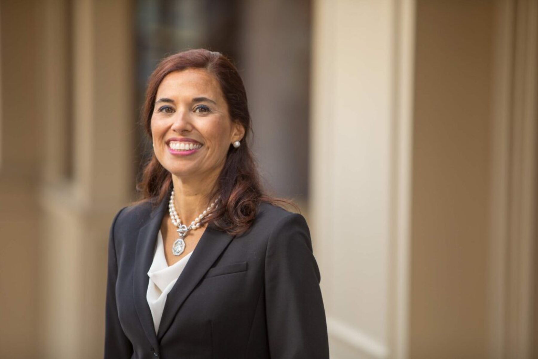 Professional executive portrait of a woman in a black blazer smiling in natural light with a soft architectural background.