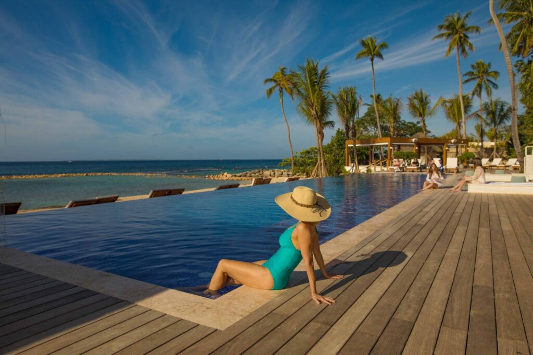 Woman relaxing by an infinity pool at a tropical beach resort under a clear blue sky.