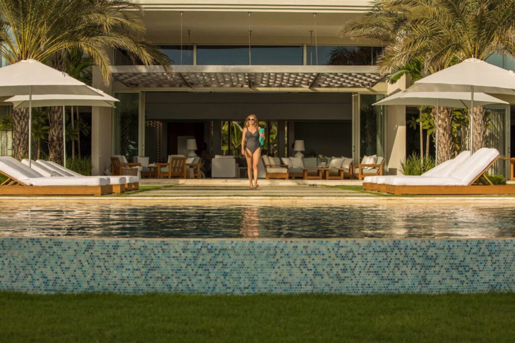 Woman walking beside a luxury resort pool in front of an open-air villa with palm trees, lounge chairs, and tropical landscaping.