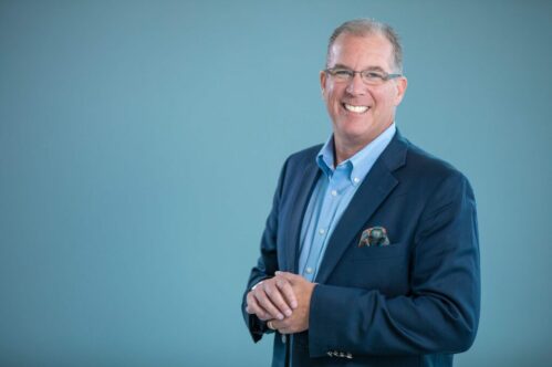 Executive Portrait – Modern Corporate Headshot Photography Professional executive portrait of a man smiling in a navy blazer and light blue shirt against a soft blue background.