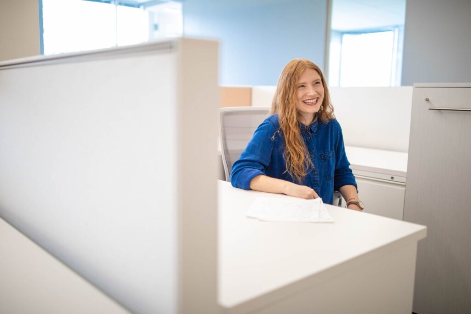 Smiling woman working in a modern office cubicle with bright natural light — workplace culture and corporate environment photography.