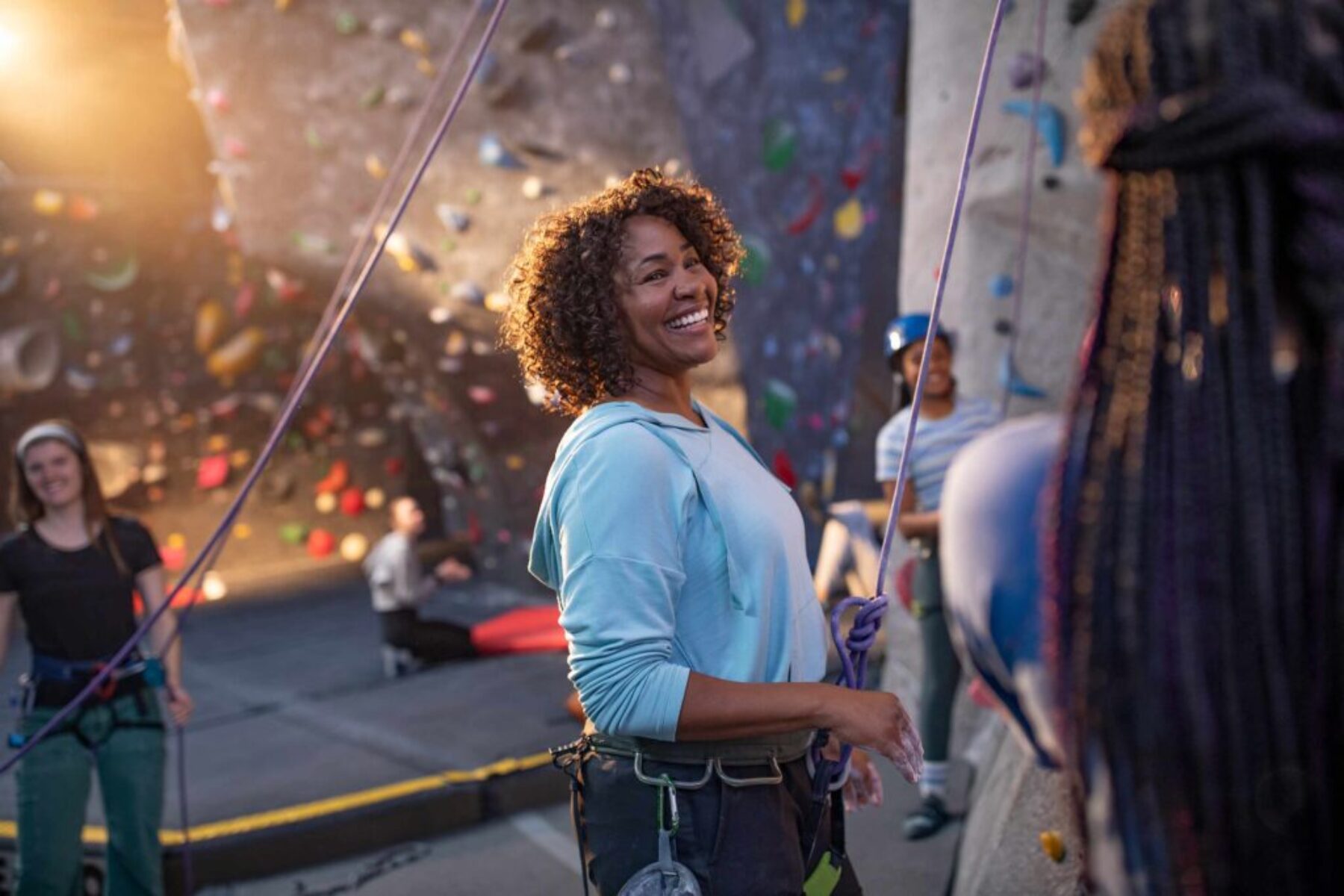 Woman smiling while rock climbing indoors with friends at a climbing gym.