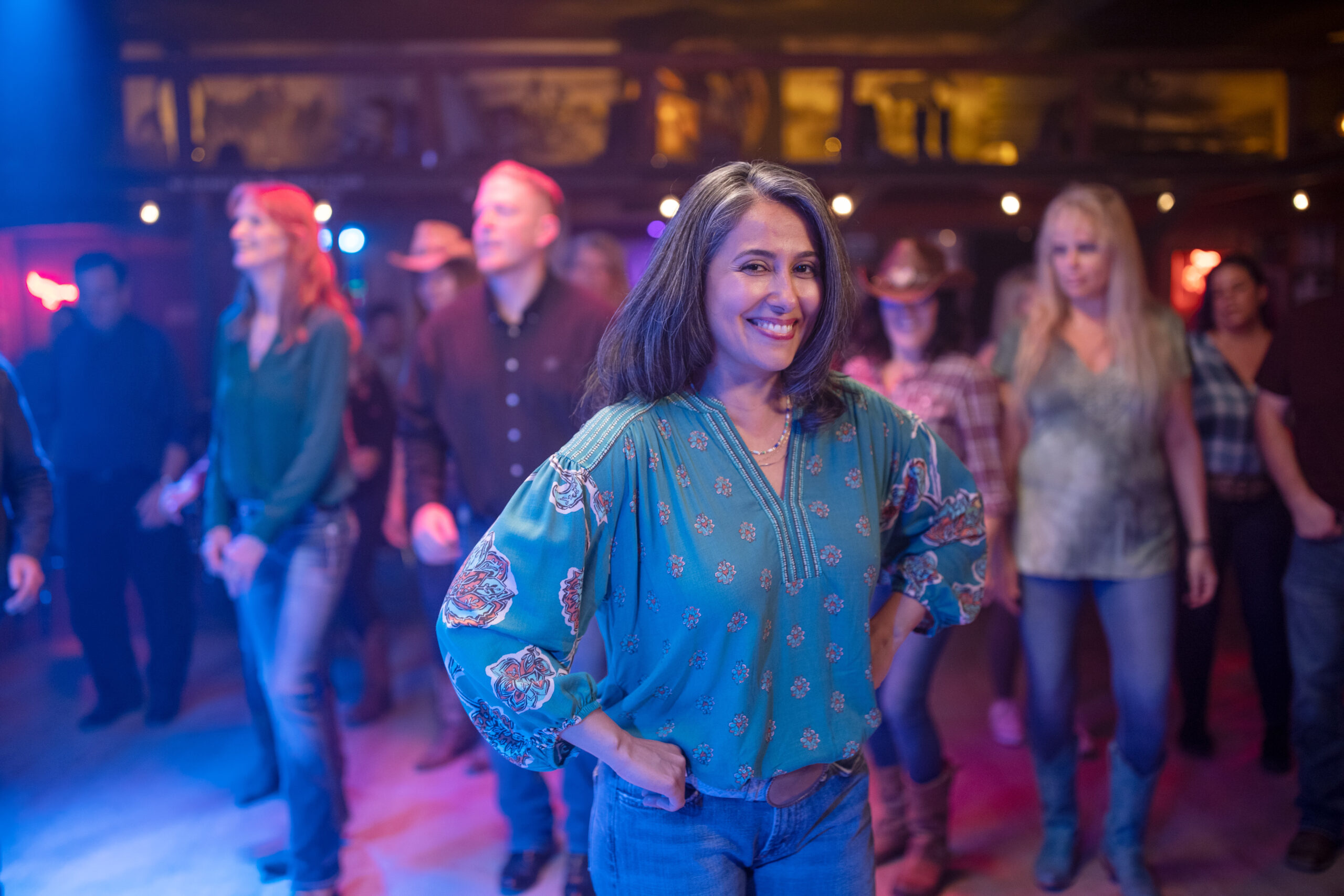 Woman smiling confidently in a dance hall with a group of people behind her during a line dancing class.