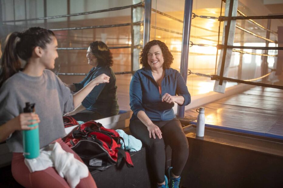 Fitness Lifestyle Photography – Post-Workout Conversation in Boxing Gym Two women smiling and talking after a workout while sitting beside a boxing ring in a gym.