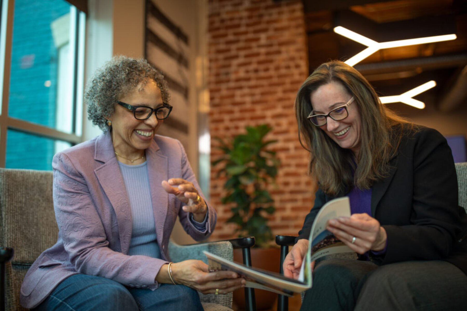 Authentic Business Branding Photography — Collaborative Meeting Portrait Branding photo of two women smiling and reviewing printed materials during a collaborative meeting in a modern office with brick and natural light.