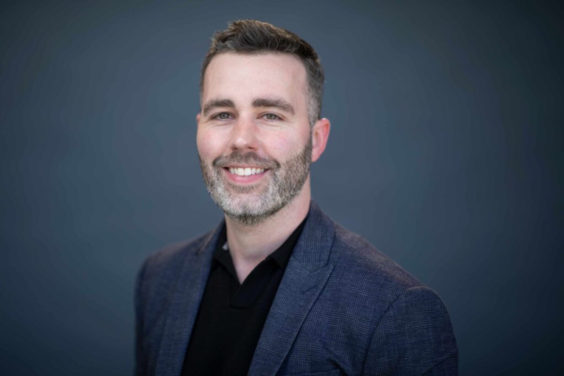 Professional executive headshot of a man smiling in a dark blazer against a soft grey background.
