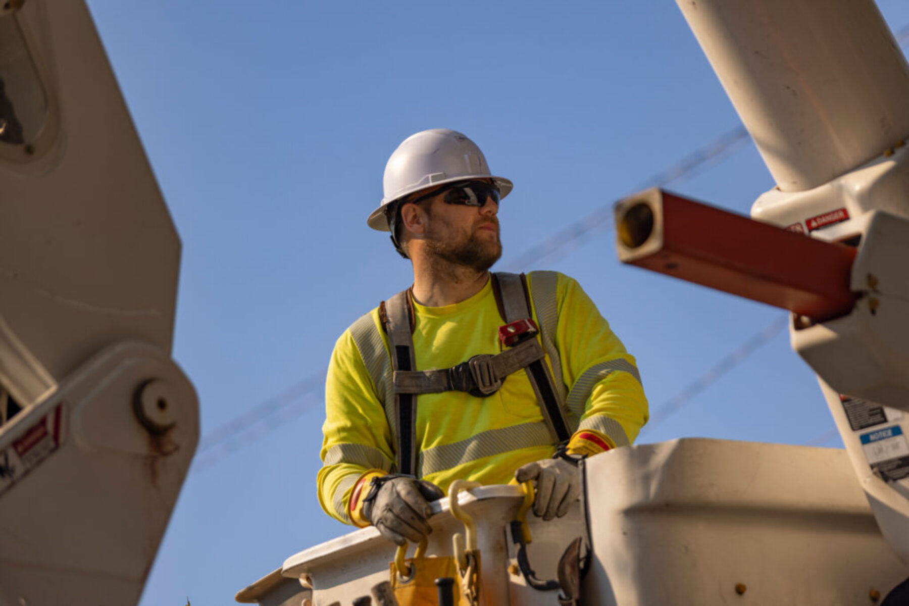utility-worker-bucket-truck-high-visibility-safety-gear-industrial-photography.jpg Utility worker in safety gear standing in a bucket truck while wearing a hard hat and high-visibility clothing.
