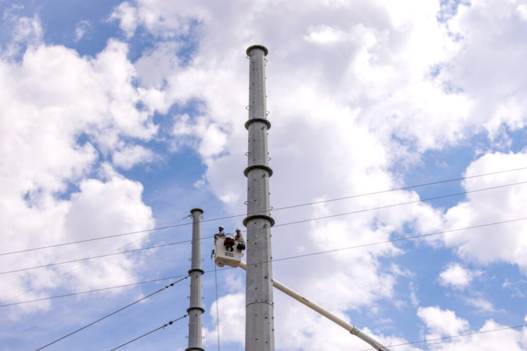 utility-workers-bucket-truck-high-voltage-lines-transmission-poles-industrial-photography.jpg Utility workers in a bucket truck working on high power lines beside tall transmission poles under a cloudy sky.