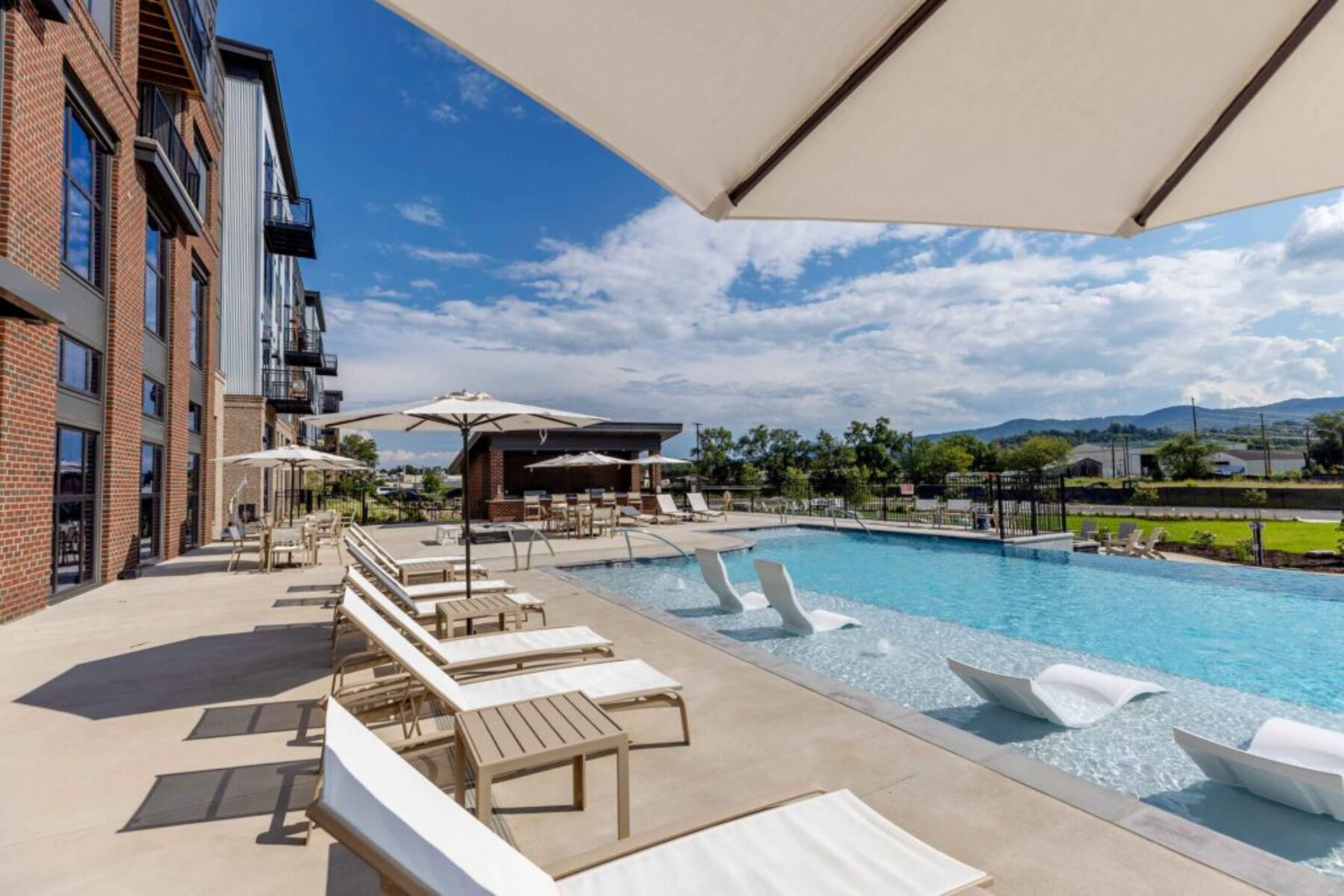 Modern apartment community pool area with lounge chairs, umbrellas, and mountain views.