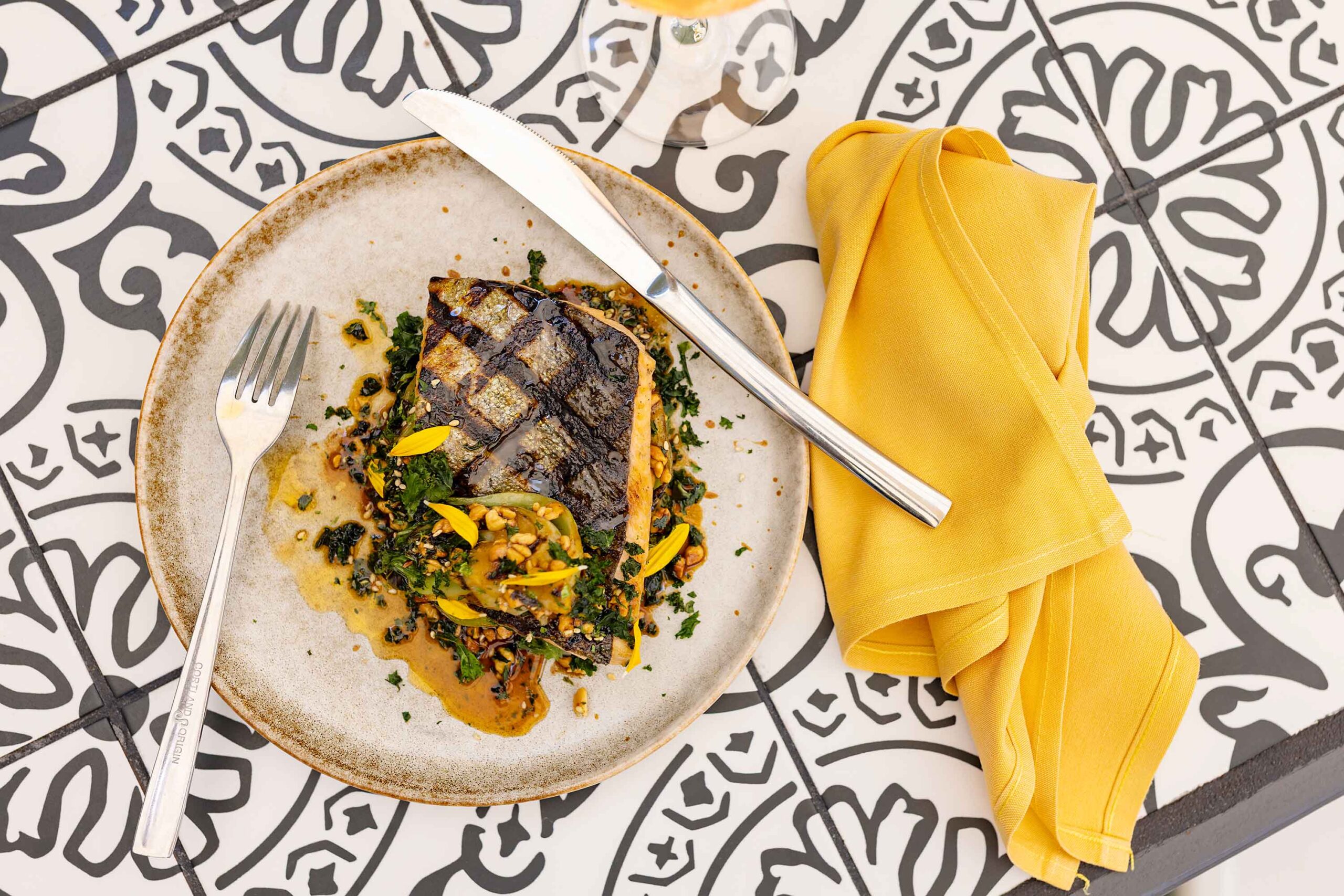 Grilled fish dish plated with vegetables and sauce on a ceramic plate, styled on patterned tiles with a yellow napkin and silver utensils.
