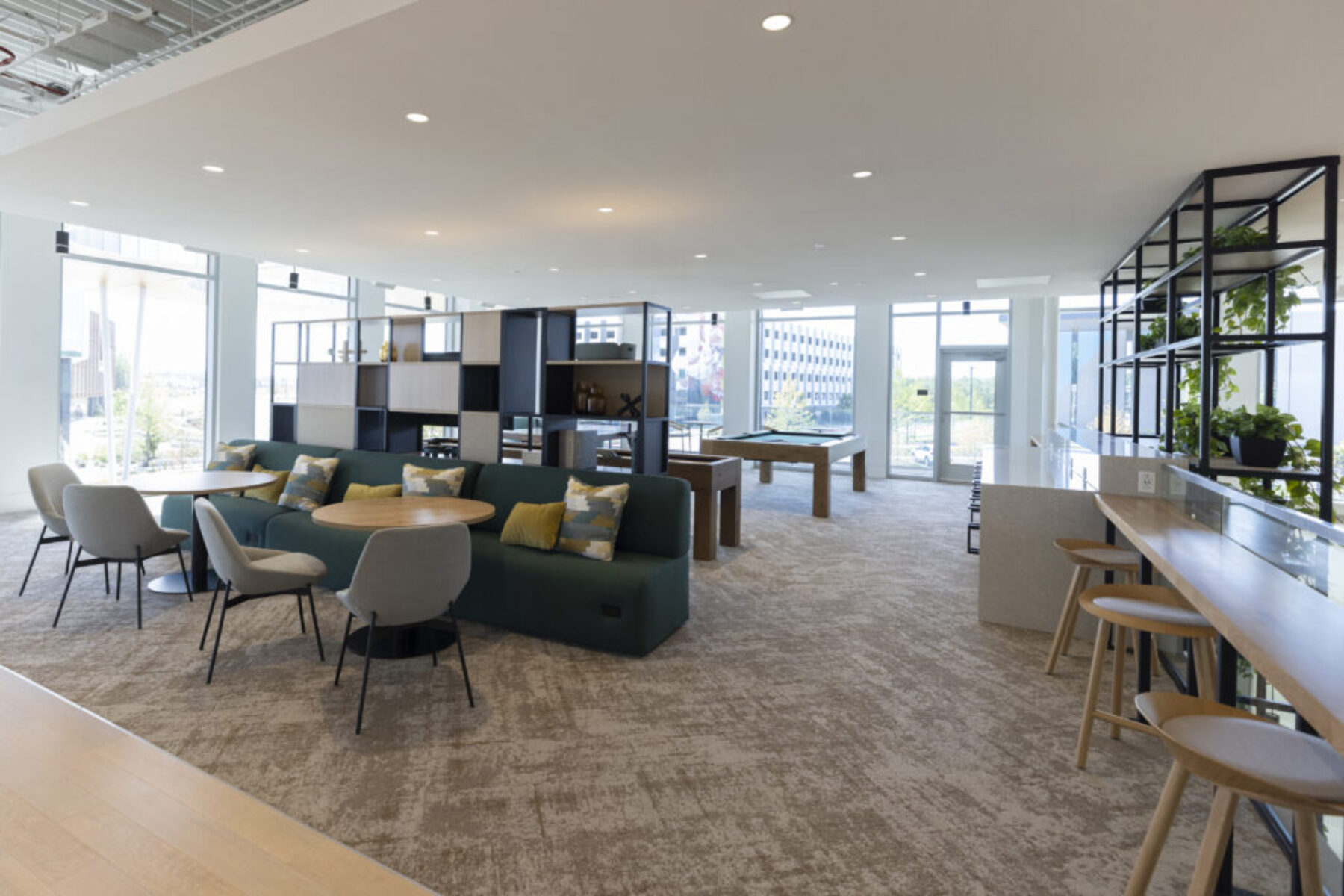 Architectural photo of a modern commercial lounge with seating areas, built-in shelving, natural light, bar-height workspace, and recreational tables in an open-concept interior.