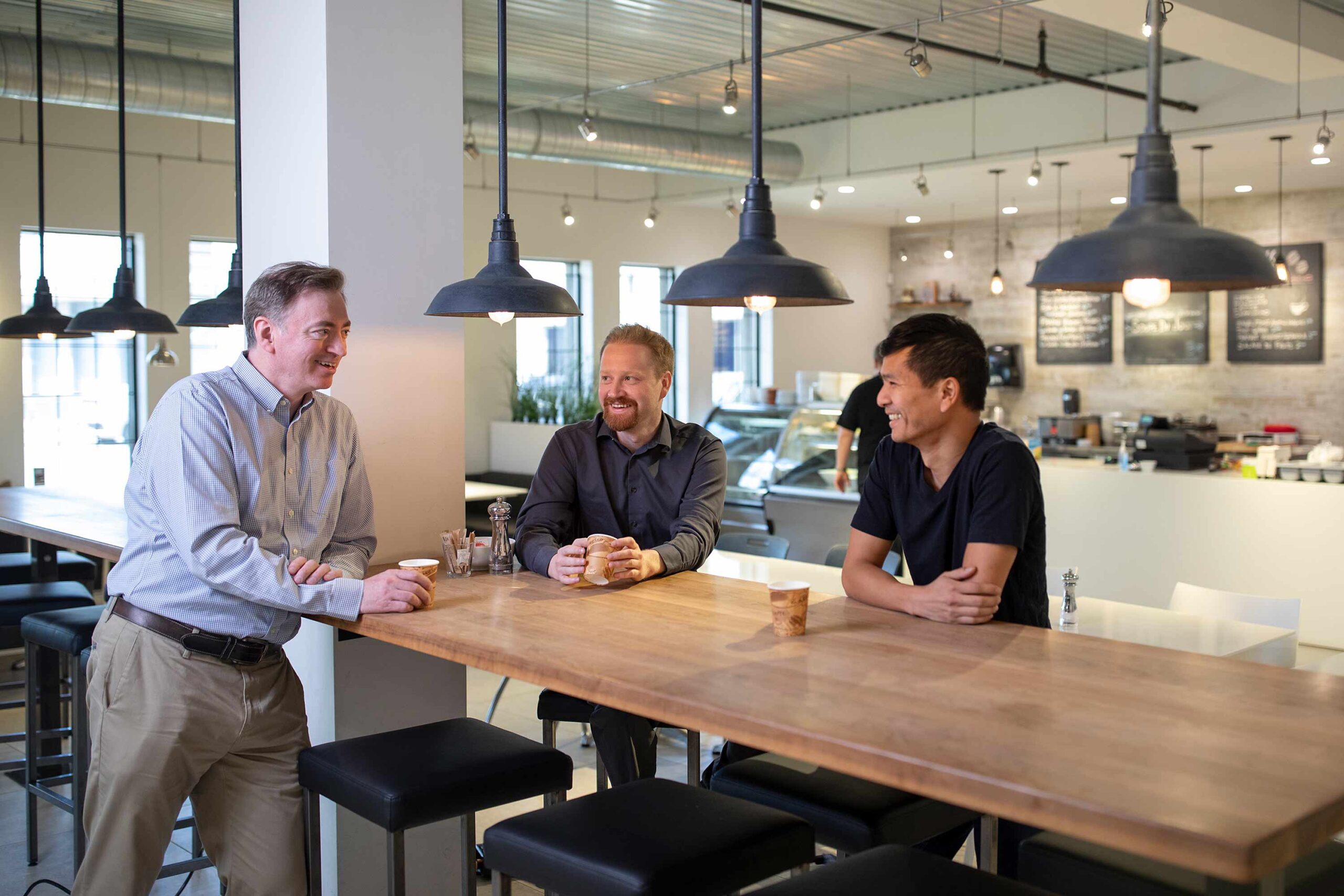 Three coworkers having a casual conversation at a coffee shop-style workplace table.