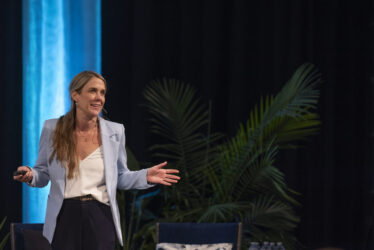A professional female speaker in a light blue blazer giving a keynote presentation on a stage with a tropical plant background.