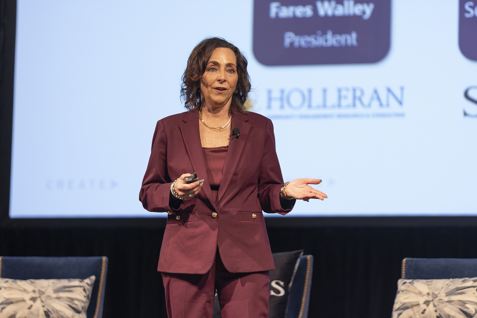 A female speaker in a maroon suit presenting on stage at a conference with a large screen behind her displaying the Holleran logo.