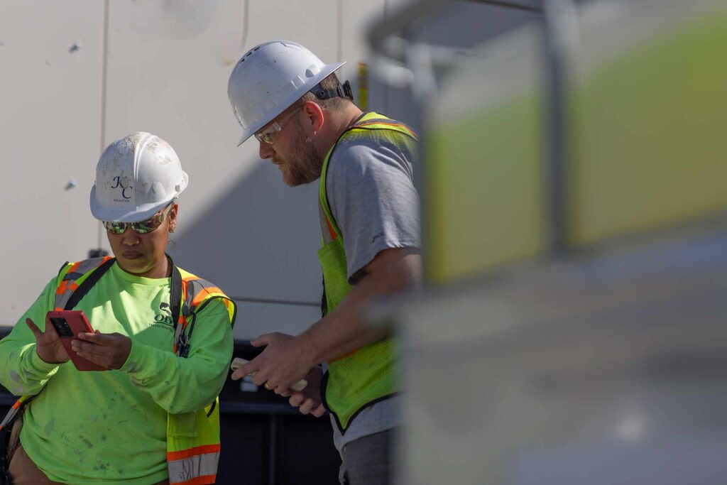 Construction workers at a project site collaborate while looking at a mobile device, using technology for safety and project tracking.