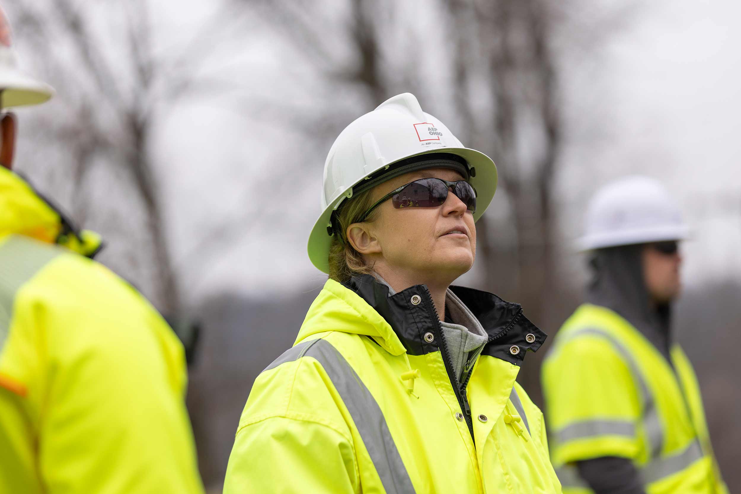 Female Appalachian Power employee wearing a white hard hat, safety glasses, and high-visibility jacket looks upward during outdoor utility operations.