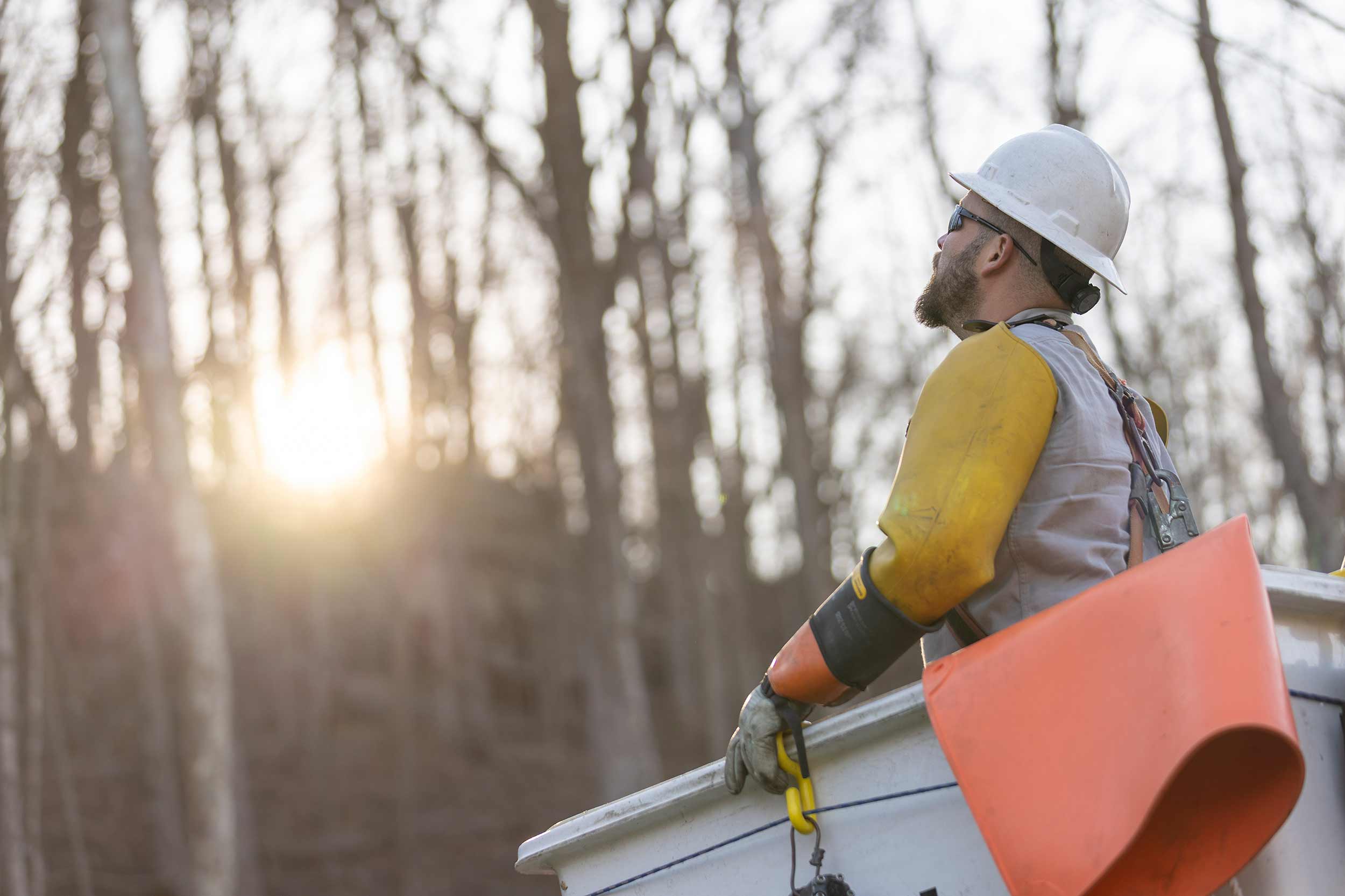 Appalachian Power lineman wearing a hard hat and protective gear looks upward from an elevated bucket truck at sunset in Virginia.