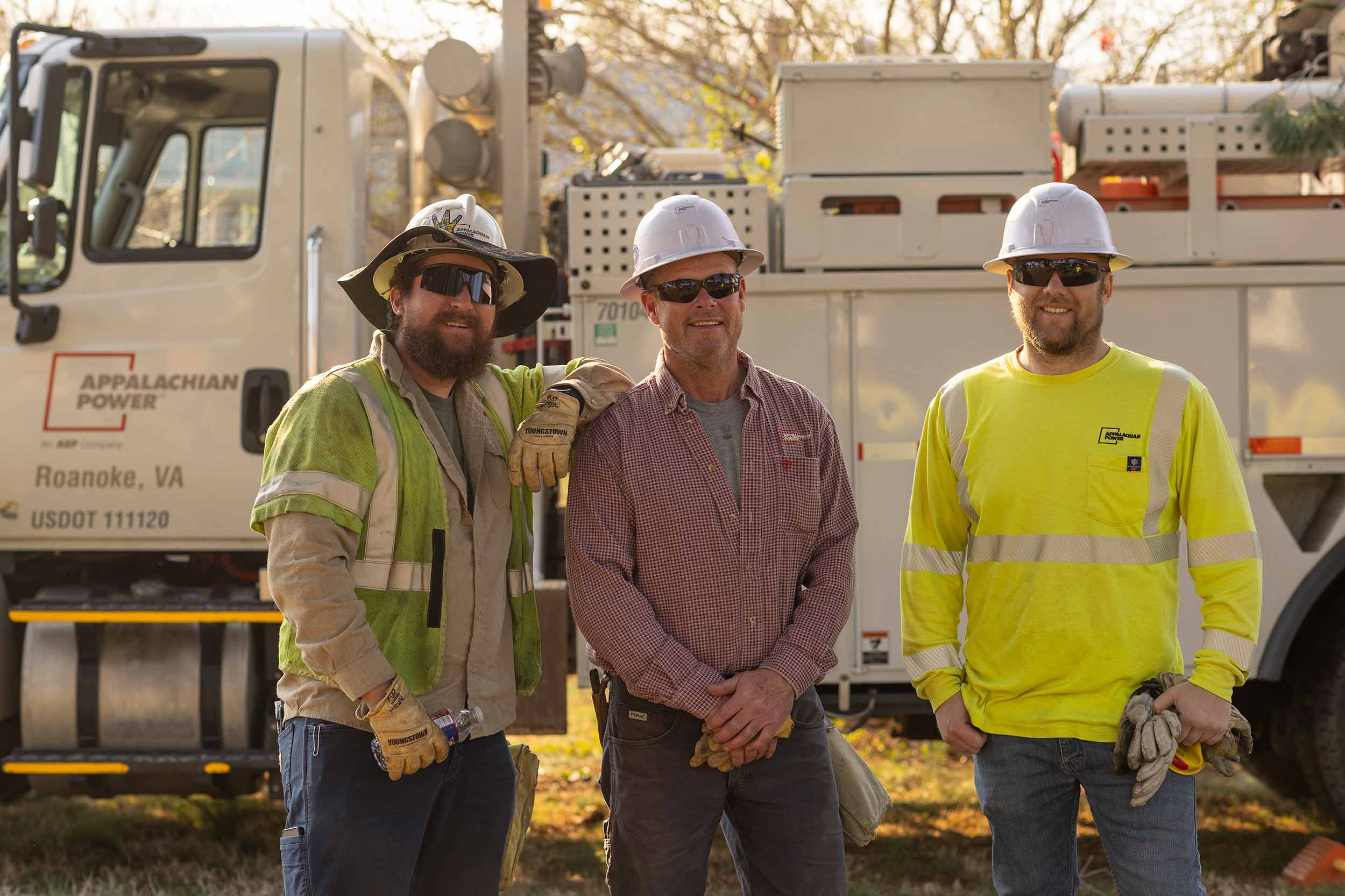 Three Appalachian Power utility crew members wearing hard hats and safety gear stand together in front of service trucks in Roanoke, Virginia.
