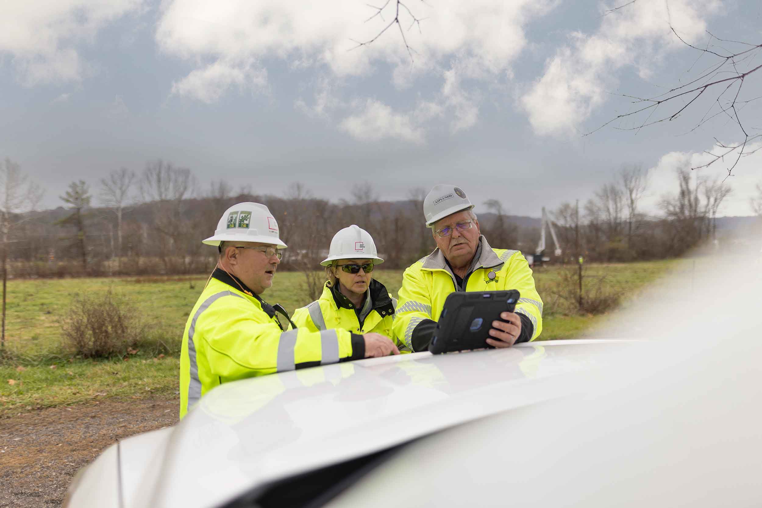 Three Appalachian Power employees wearing hard hats and high-visibility safety jackets review information on a tablet at an outdoor worksite in Virginia.