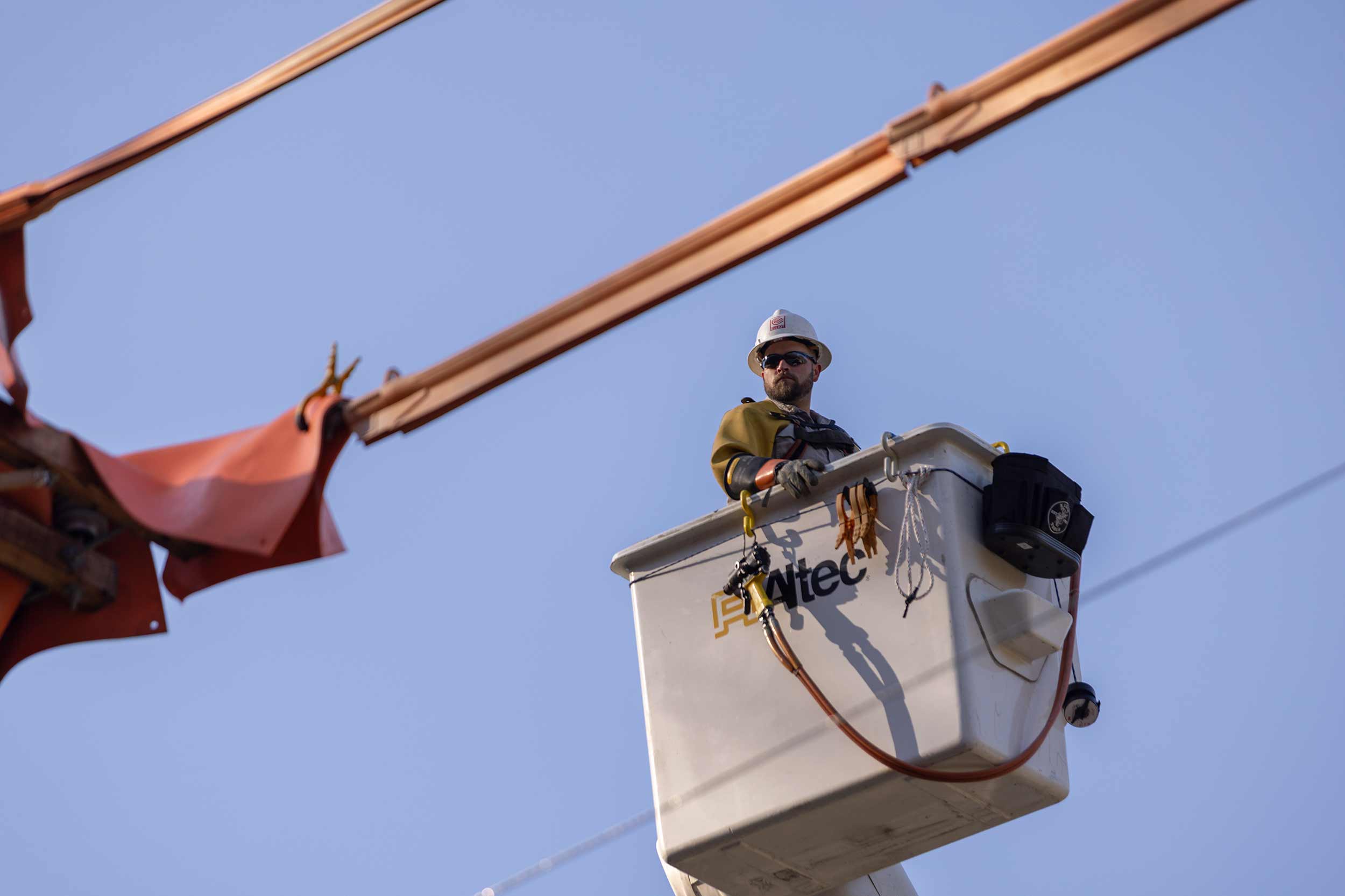 Utility lineman wearing a hard hat and safety equipment stands in an elevated bucket truck beside overhead electrical lines against a blue sky.